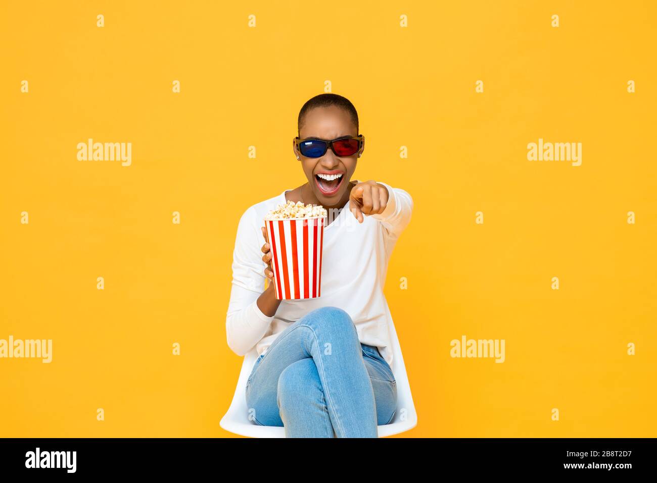 Fröhliche Amerikanerin mit 3D-Brille und Popcorn lacht, während sie den Film auf gelbem Studiohintergrund sieht Stockfoto
