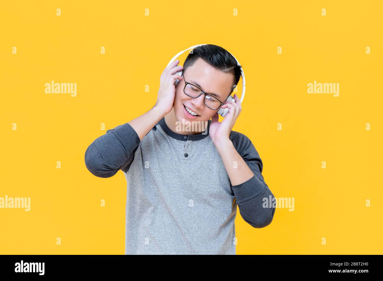 Happy Asian man mit Kopfhörern, die entspannende Musik hören, mit geschlossenen Augen auf gelbem Hintergrund Stockfoto
