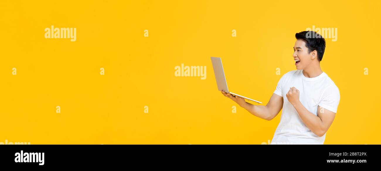 Glücklicher junger Asian, der einen Laptop mit sich führt und seine Faust mit Ja hebt, indem er auf gelbem Bannerhintergrund mit Platz für Kopien isoliert ist Stockfoto