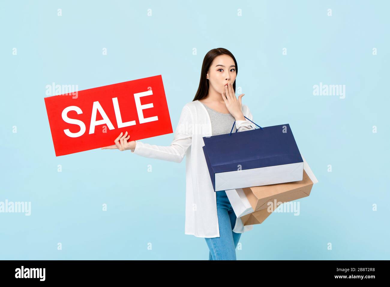 Überraschte die schöne asiatische Frau mit Einkaufstaschen, die ein rotes Verkaufszeichen auf hellblauem Hintergrund zeigen Stockfoto
