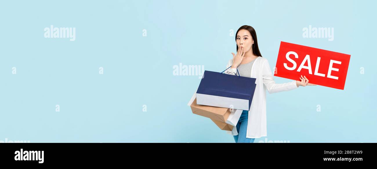Überraschte die schöne asiatische Frau mit Einkaufstaschen, die ein rotes Verkaufszeichen auf hellblauem Bannerhintergrund mit Kopierraum zeigt Stockfoto