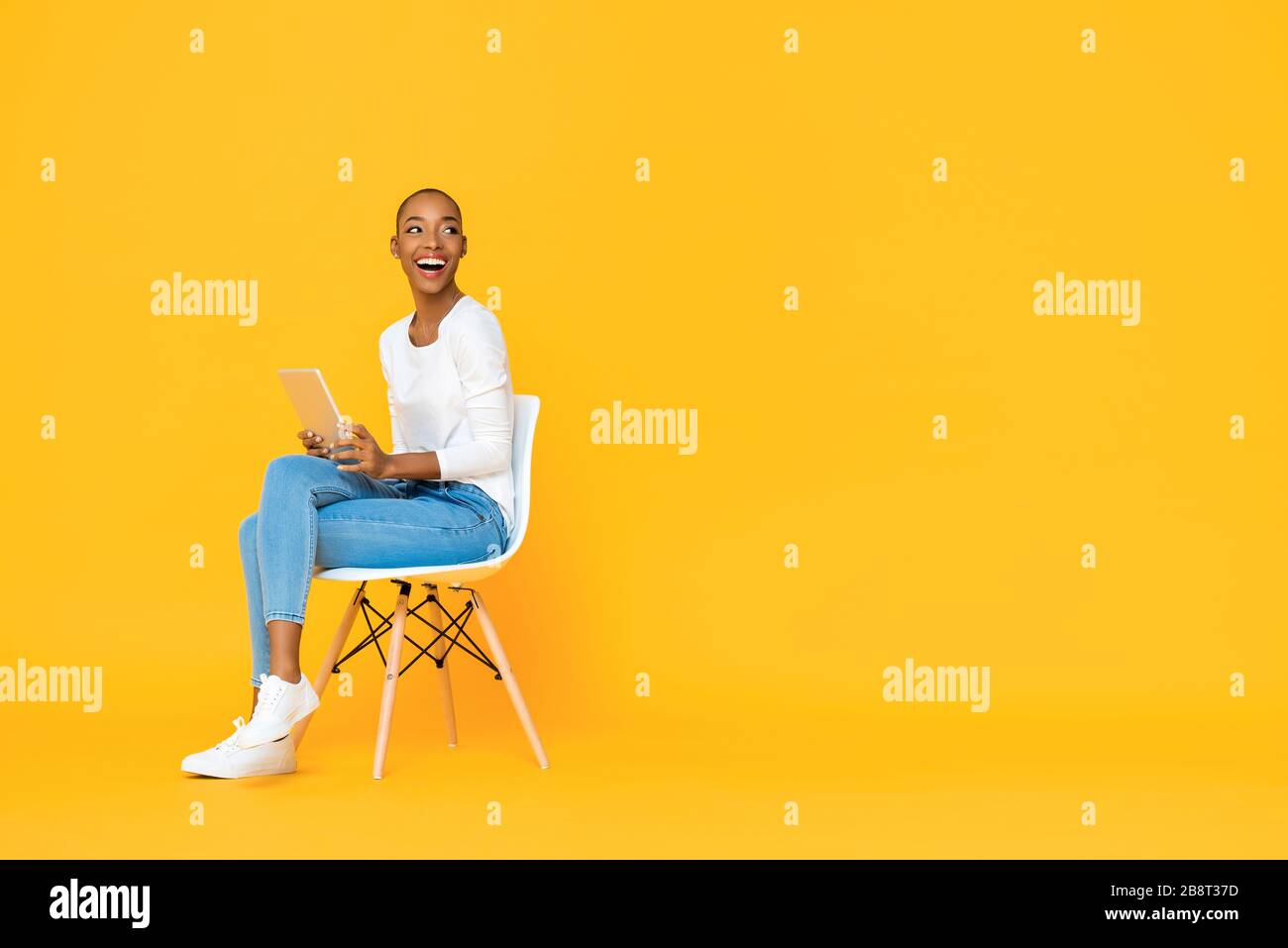 Trendige, lächelnde afroamerikanische Frau, die mit einem Tablet-Computer auf einem Stuhl sitzt und auf leeren Raum neben isoliertem gelbem Hintergrund schaut Stockfoto