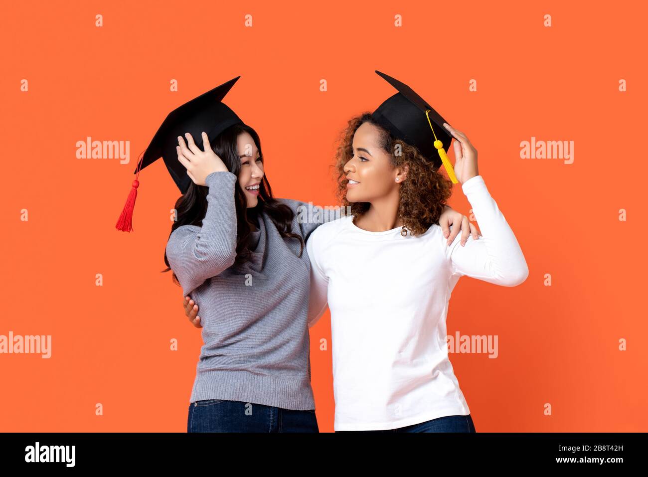 Porträt zweier glücklicher Mixed-Race-Frauen, die dort Graduierung Caps halten und sich gegenseitig in orange isoliertem Studiohintergrund betrachten Stockfoto