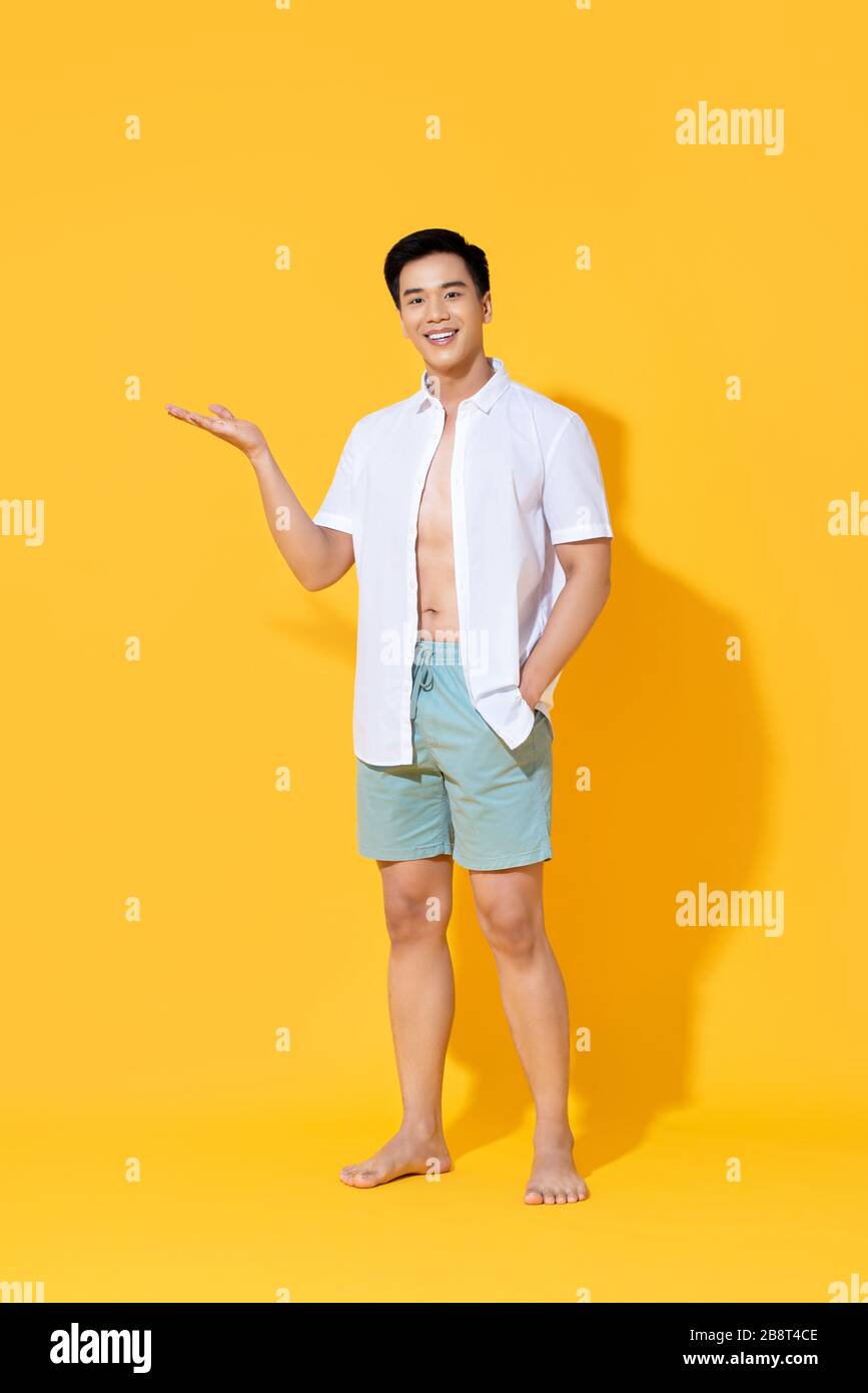 Junge lächelnde, gutaussehende Asiaten in Strandkleidung, die eine offene Handbewegung ausführen und im Sommer gelb isolierte Studiohintergrund unbeschriftet Platz zeigen Stockfoto