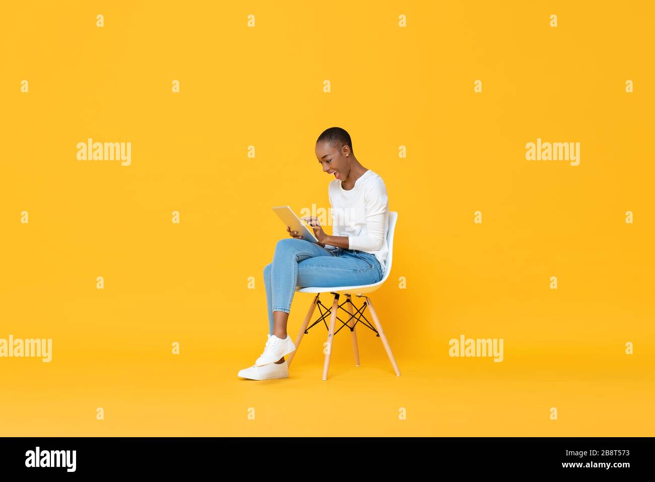 Glückliche junge afroamerikanische Frau, die mit einem Tablet-Computer in gelb isoliertem Studiohintergrund auf einem Stuhl sitzt Stockfoto
