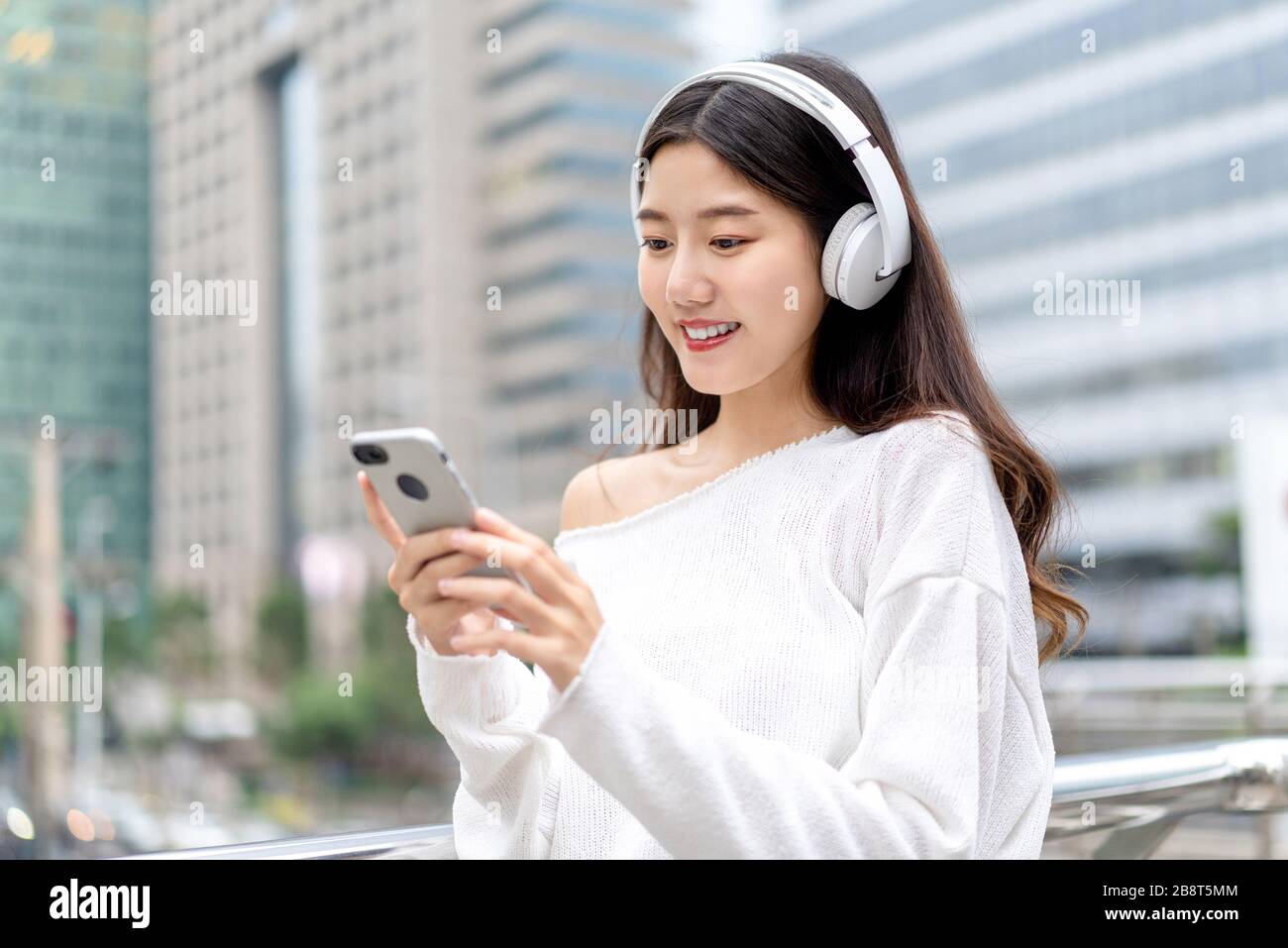 Junges asiatisches Mädchen, das Kopfhörer trägt, um Musik vom Handy gegen den Backround des Stadtgebäudes zu hören Stockfoto