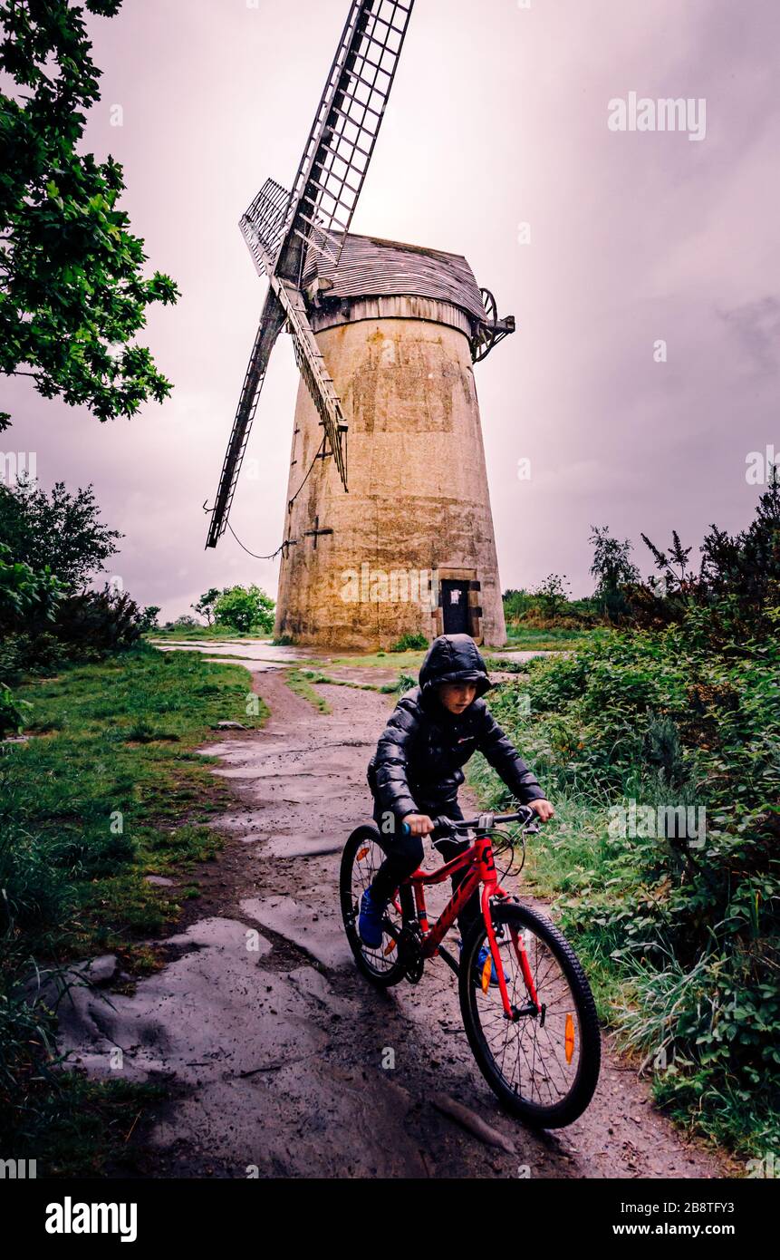 Der Junge, der mit dem roten Fahrrad im Regen unterwegs ist, passiert die alte historische Windmühle Bidston Hill Wirral UK Stockfoto