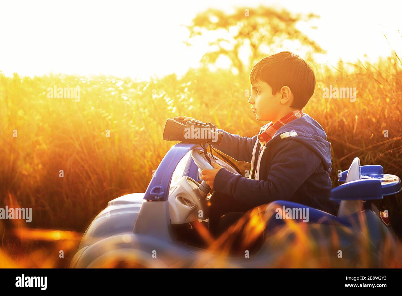 Ein Kind, das mitten auf dem Land sitzt und bei Sonnenuntergang auf seinem Spielzeugauto sitzt, sieht ernst aus Stockfoto