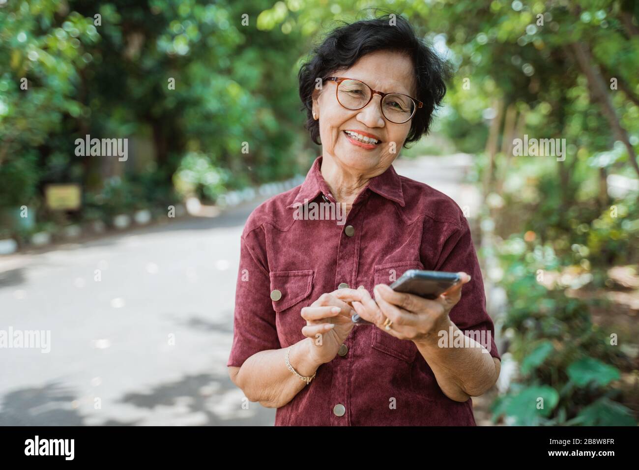 Lächelnde asiatische alte Frau, die ein Handy benutzt, wenn sie zur Kamera schaut Stockfoto