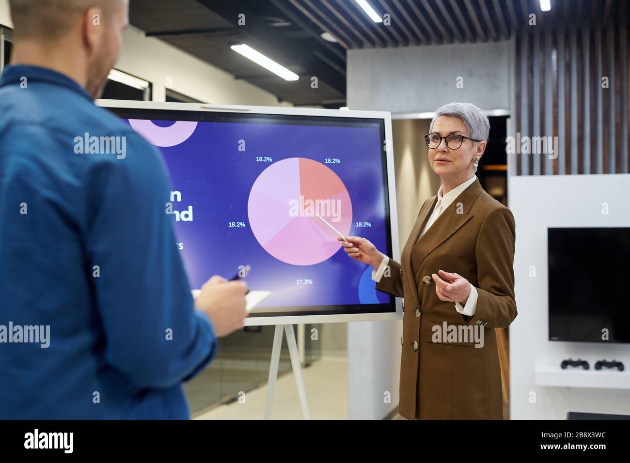 Portrait der modernen reifen Geschäftsfrau, die auf ein Tortendiagramm zeigt, während sie Mitarbeitern oder Kunden in der Bürohalle eine Präsentation vorgibt Stockfoto