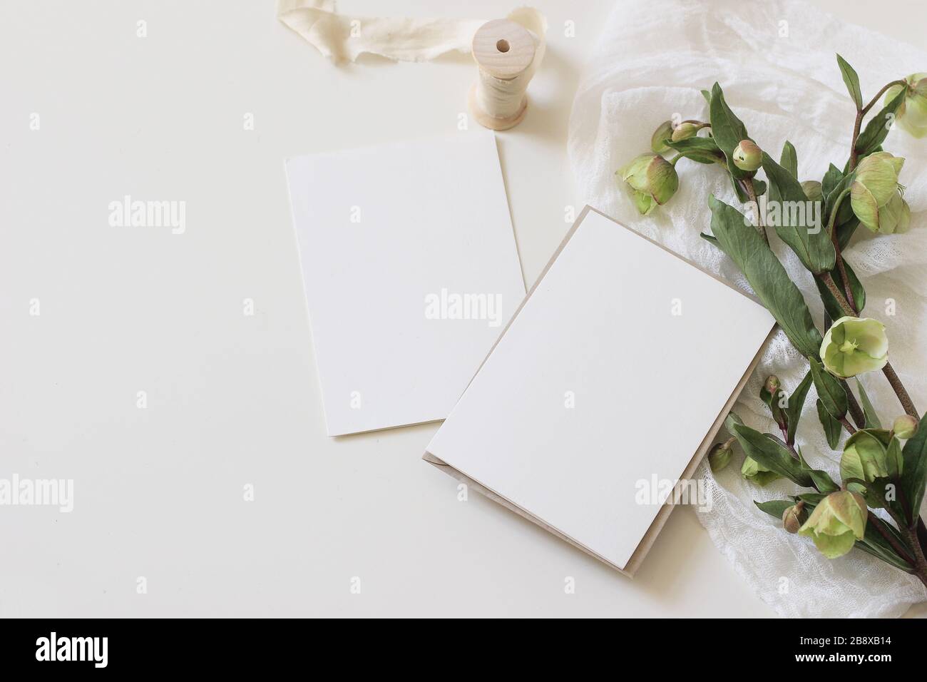 Stockfoto im Stil eines Hochzeitsfrühlings. Feminine Desktop-Mockup-Szene mit grünen Hellebores Blumen, Seidenband, Muslin-Tuch und leerem Papier-Grußwagen Stockfoto