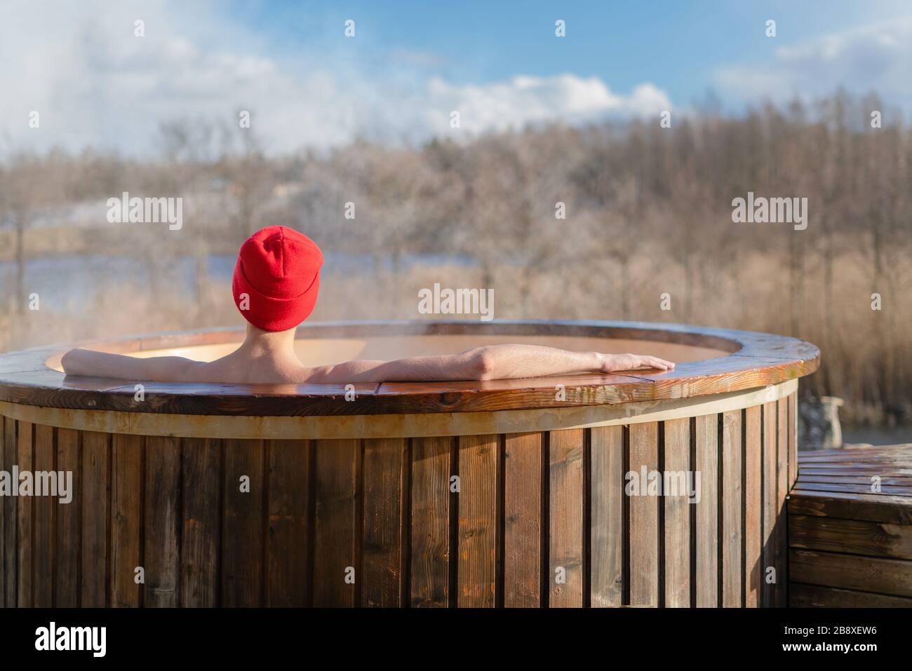 Junger Erwachsener, der draußen im Wannen-Whirlpool aus Holz entspannen und die Natur betrachten kann. Person, die an einem sonnigen Tag einen heißen Dampfpool genießen, private Spa-Behandlung Stockfoto