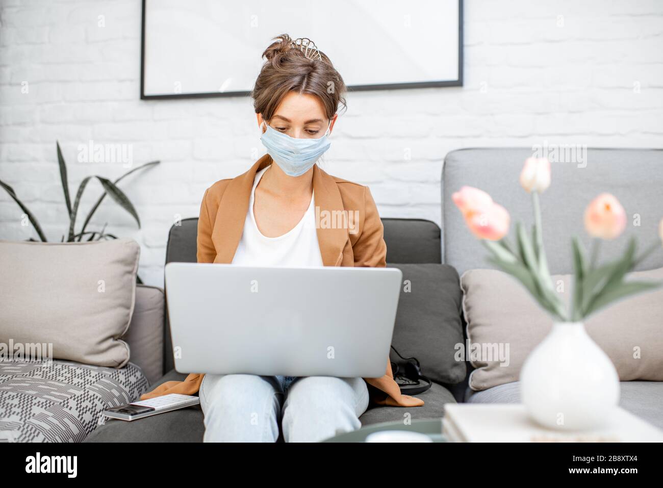 Frau in medizinischer Maske, die online von zu Hause aus auf dem Laptop arbeitet. Bleiben Sie während eines Virenpandemic-Konzepts zu Hause Stockfoto