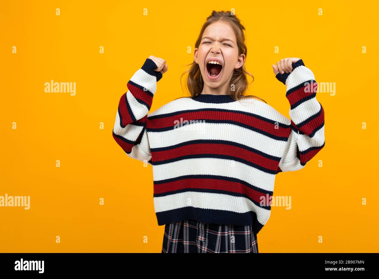 Teenager-Mädchen in einem gestreiften Pullover mit weit geöffnetem Mund und erhobenen Armen ruft die Nachrichten auf gelbem Studiohintergrund mit Kopierraum auf Stockfoto