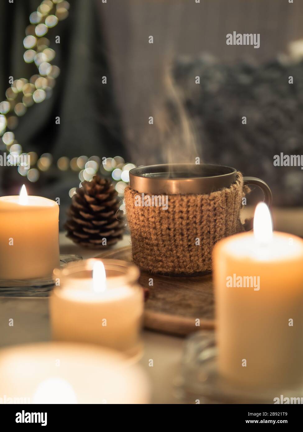 Gemütliches Zuhause, Hyge, Gemütlichkeit - heißes Getränk in der Tasse mit Dampf und brennenden weißen Duftkerzen in der Nähe des Sofas mit Kissen und Plaid. Winterdekor und heiße Teetasse. Vertikal Stockfoto