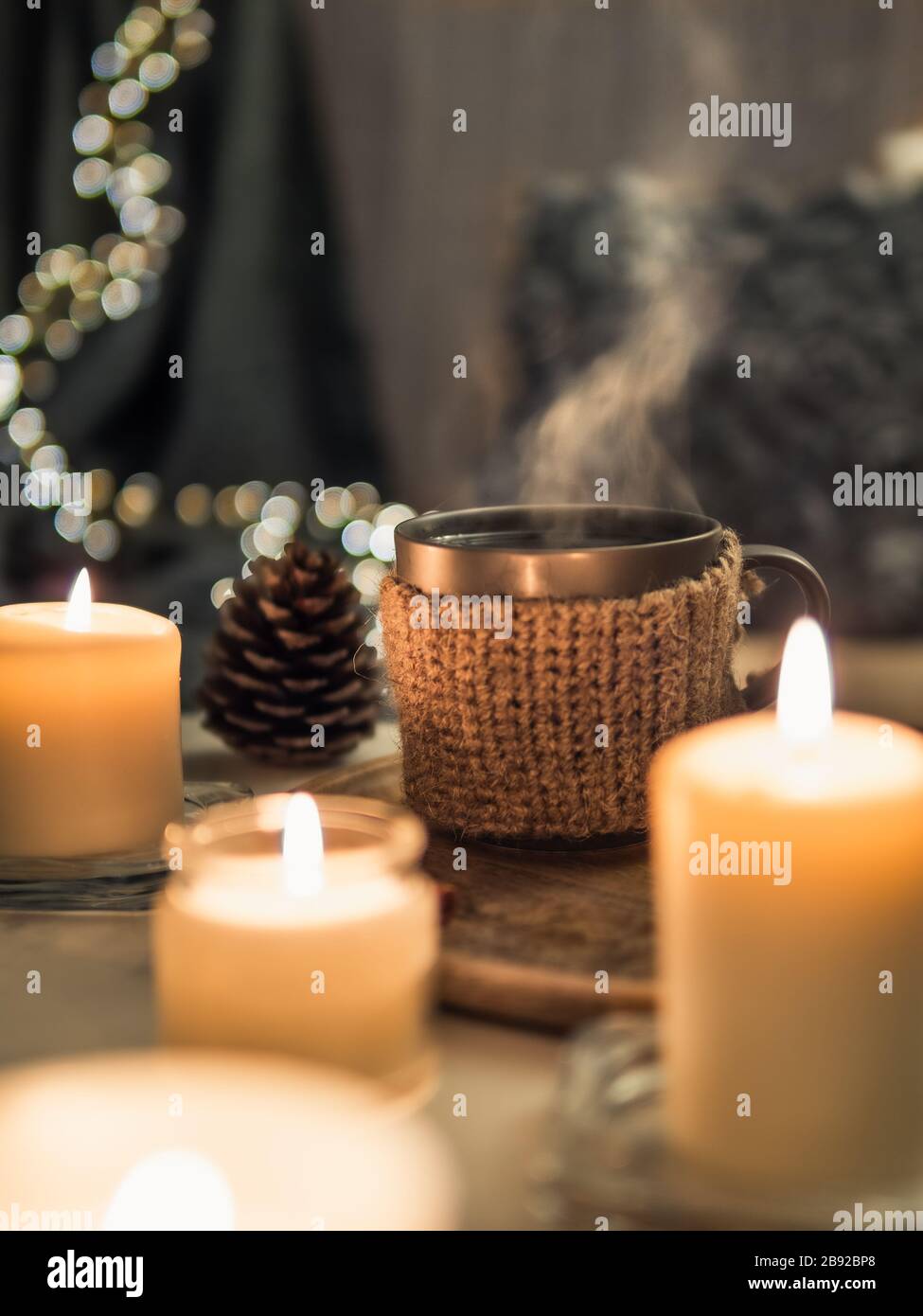 Gemütliches Zuhause, Hyge, Gemütlichkeit - heißes Getränk in der Tasse mit Dampf und brennenden weißen Duftkerzen in der Nähe des Sofas mit Kissen und Plaid. Winterdekor und heiße Teetasse. Vertikal Stockfoto