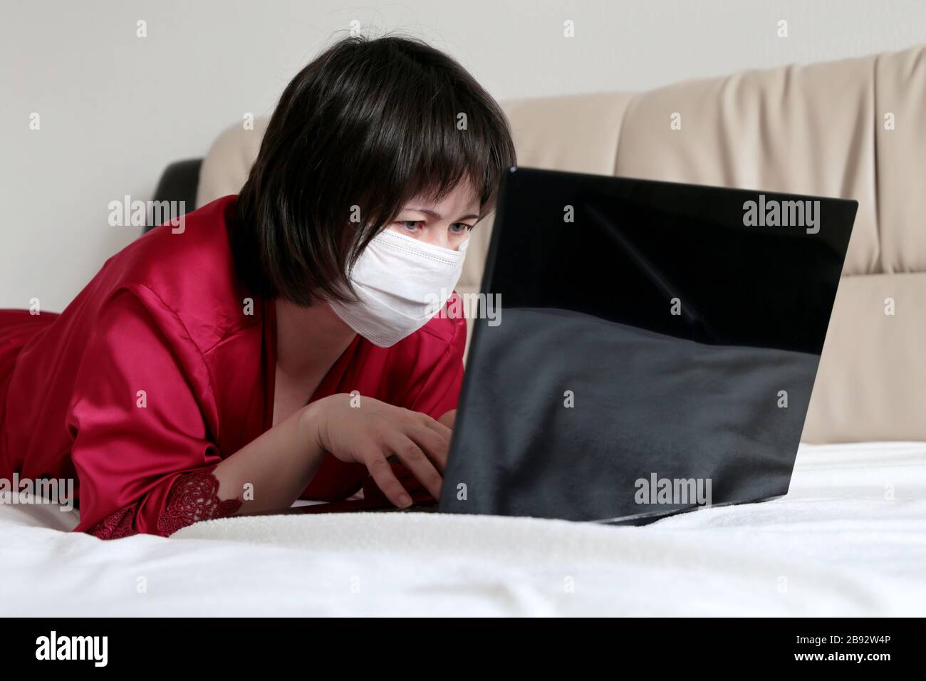 Frau in medizinischer Gesichtsmaske, die mit einem Laptop auf einem Bett liegt. Konzept der Remotearbeit während einer COVID-19-Coronavirus-Epidemie, Hausquarantäne Stockfoto