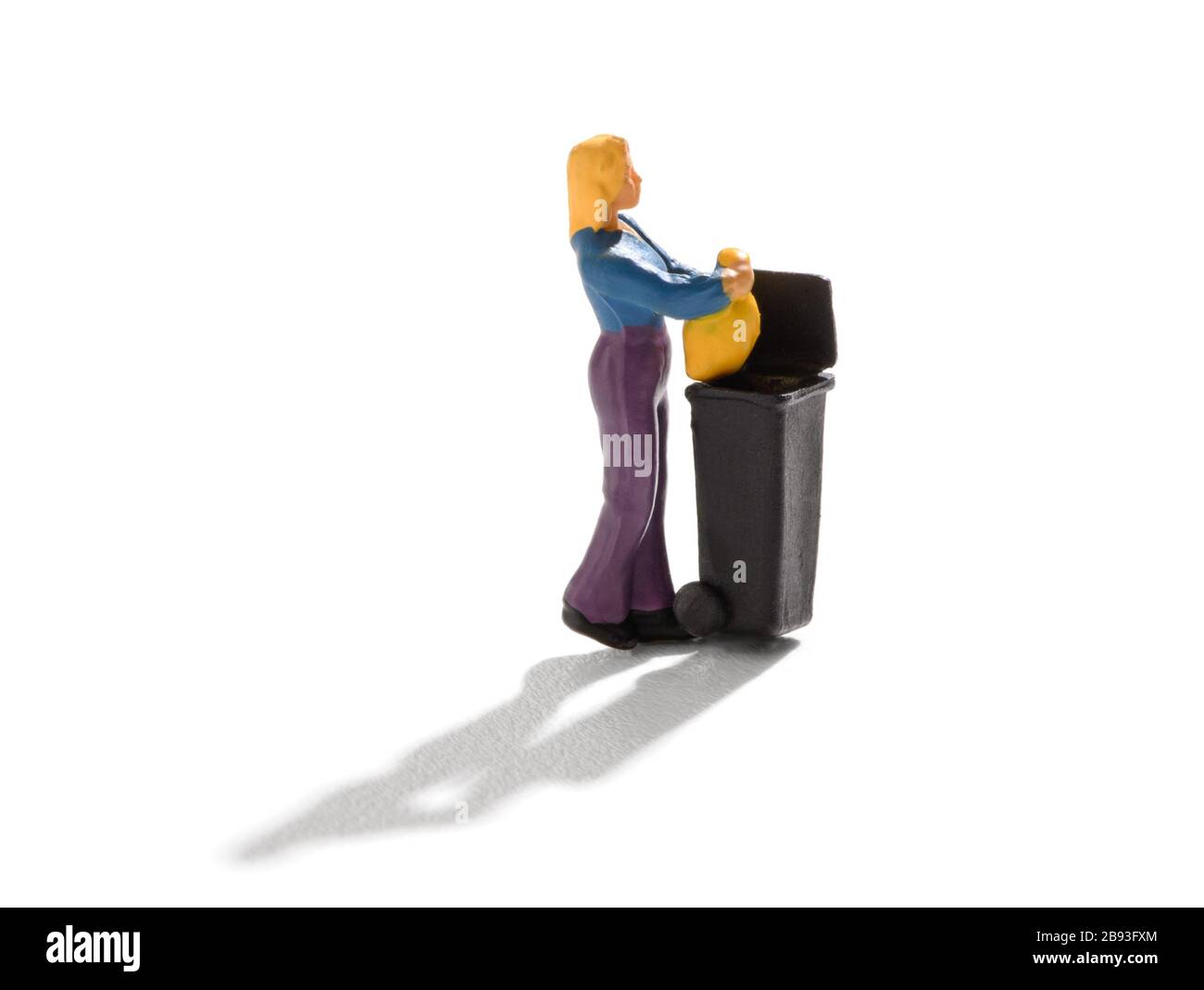 Miniaturfrau wirft den Hausmüll aus und legt ihn in einem Abfallentsorgungskonzept auf weiß mit Schatten in einen großen schwarzen Mülleimer Stockfoto