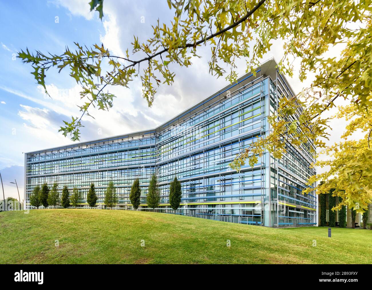 Außenansicht eines modernen Bürogebäudes mit eckiger Glasfront in einer Quelllandschaft mit grünen Rasenflächen und Bäumen unter blauem bewölktem Himmel Stockfoto