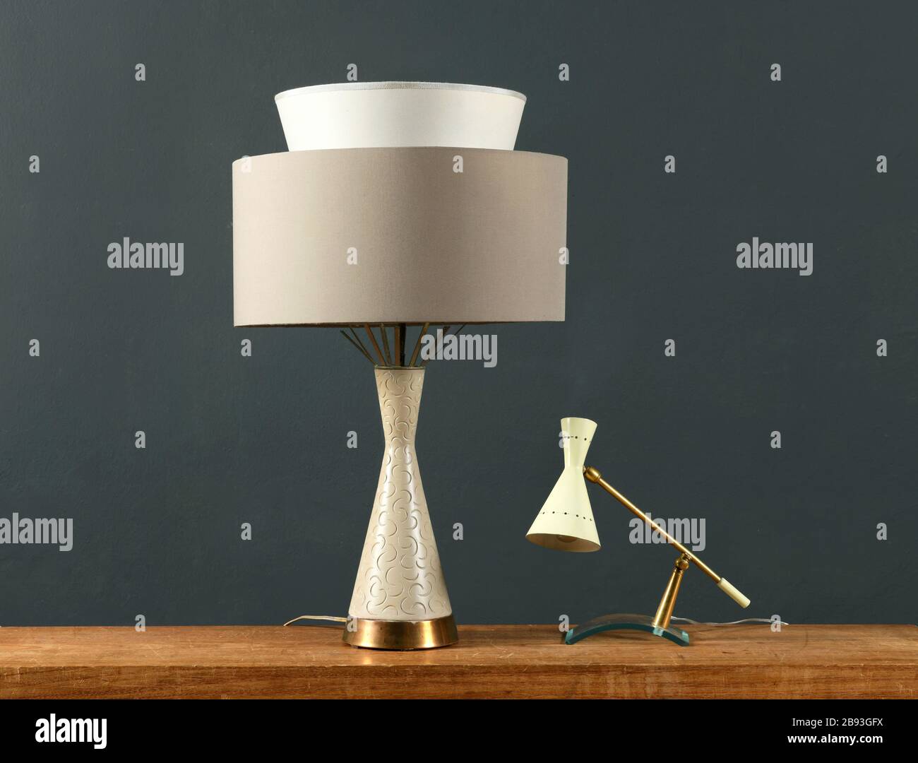 Zwei Vintage-Lampen an einem Holzschrank, eine zylindrische mit eckigem Boden und Schatten und eine kleine Anglepoise-Lampe gegen eine dunkelgraue Wand Stockfoto