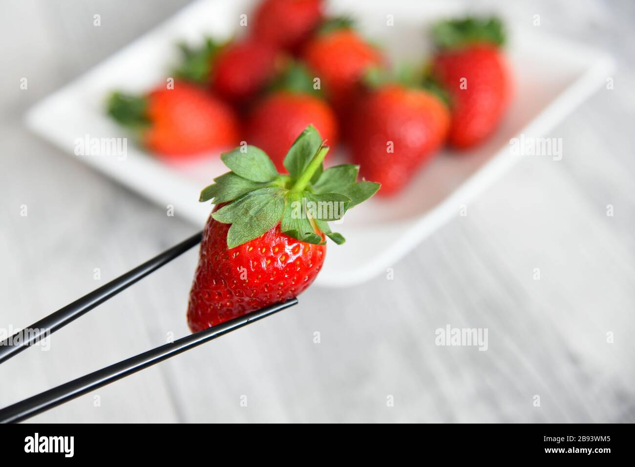 Nahaufnahme von Essstäbchen, die eine rote saftige Erdbeere mit grünen Blättern vor einem verschwommenen Hintergrund halten. Gesunde organische Erdbeeren. Stockfoto