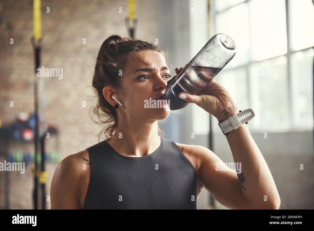 Nahaufnahme der jungen Sportfrau in Ohrhörern ist das Trinkwasser beim Training im Fitnessstudio. Profisport. TRX-Schulung. Gesunder Lebensstil Stockfoto