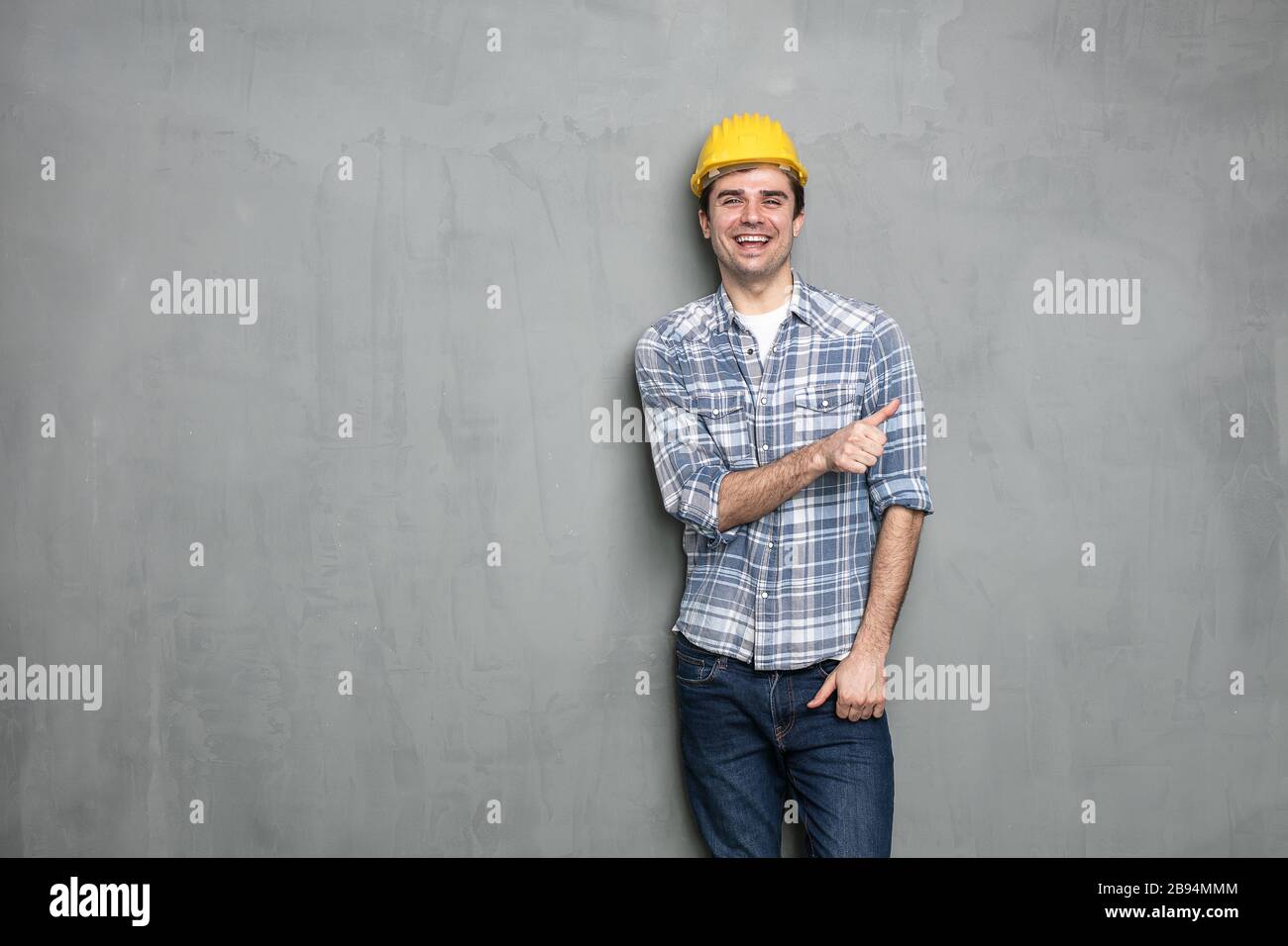 Selbstbewusster junger Arbeiter mit Sicherheitshelm, der eine leere Seite des Bildes für Text oder Werbung für etwas darstellt, an einer betongrauen Wand Stockfoto