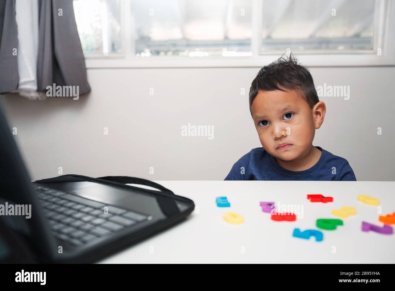 Ein männliches Kind, das wenig Interesse am Lehrplan der Homeschule zeigt, direkt vor einem Laptop sitzt und Manipulative anführt. Stockfoto