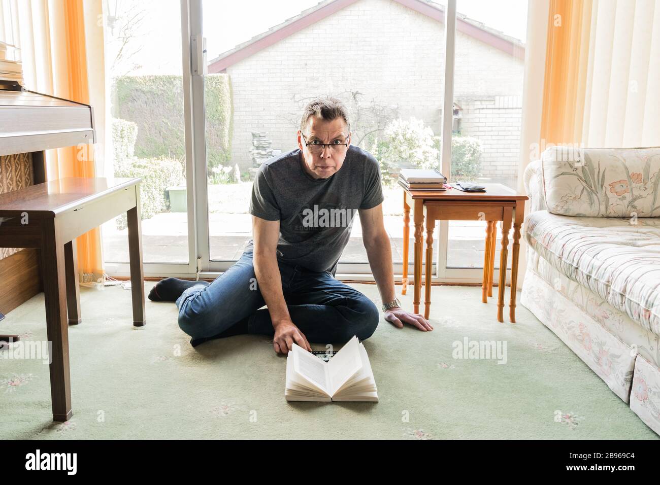 50 etwas männlich in Coronavirus Selbstisolierung zu Hause, sitzen vor dem Fenster im Wohnzimmer und lesen ein Buch Stockfoto