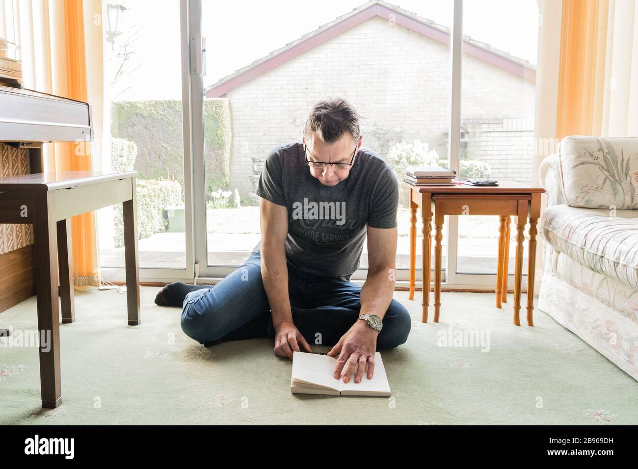 50 etwas männlich in Coronavirus Selbstisolierung zu Hause, sitzen vor dem Fenster im Wohnzimmer und lesen ein Buch Stockfoto