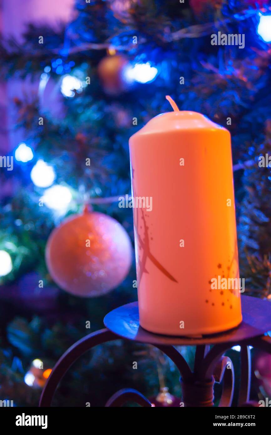 Eine große orangefarbene Kerze auf einem schmiedeeisernen Standfuß vor dem Hintergrund eines mit Spielzeug verzierten Weihnachtsbaums ist ein Detail des Innenraums. Vertikales Foto. Neujahrsfeiertag verschwommene Hintergrundseite. Stockfoto