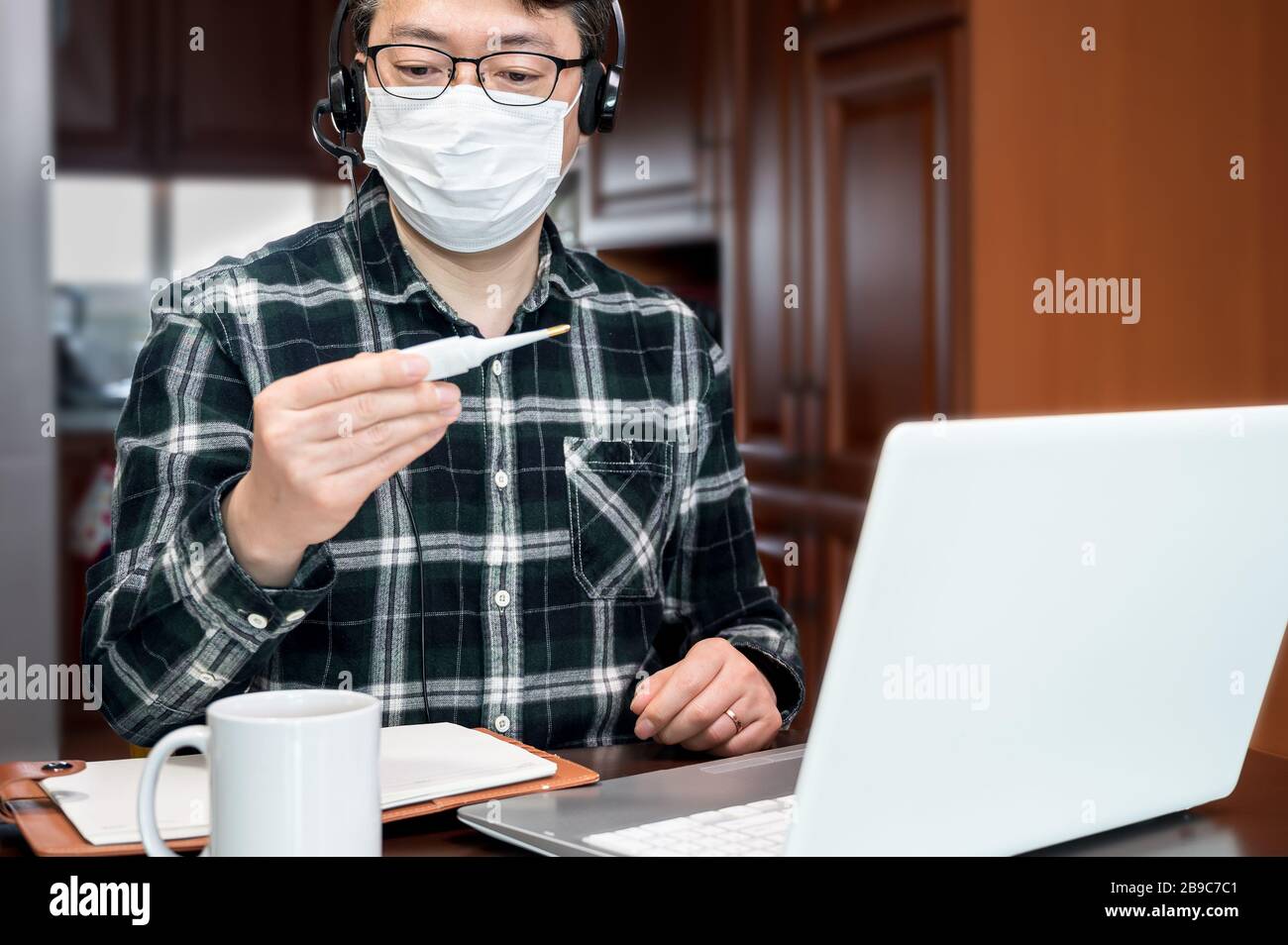 Ein asiatischer Mann, der aufgrund einer massiven Pandemie selbst isoliert und von zu Hause aus arbeitet. Ein Mann, der ein Thermometer hält, um die Körpertemperatur zu messen. Stockfoto