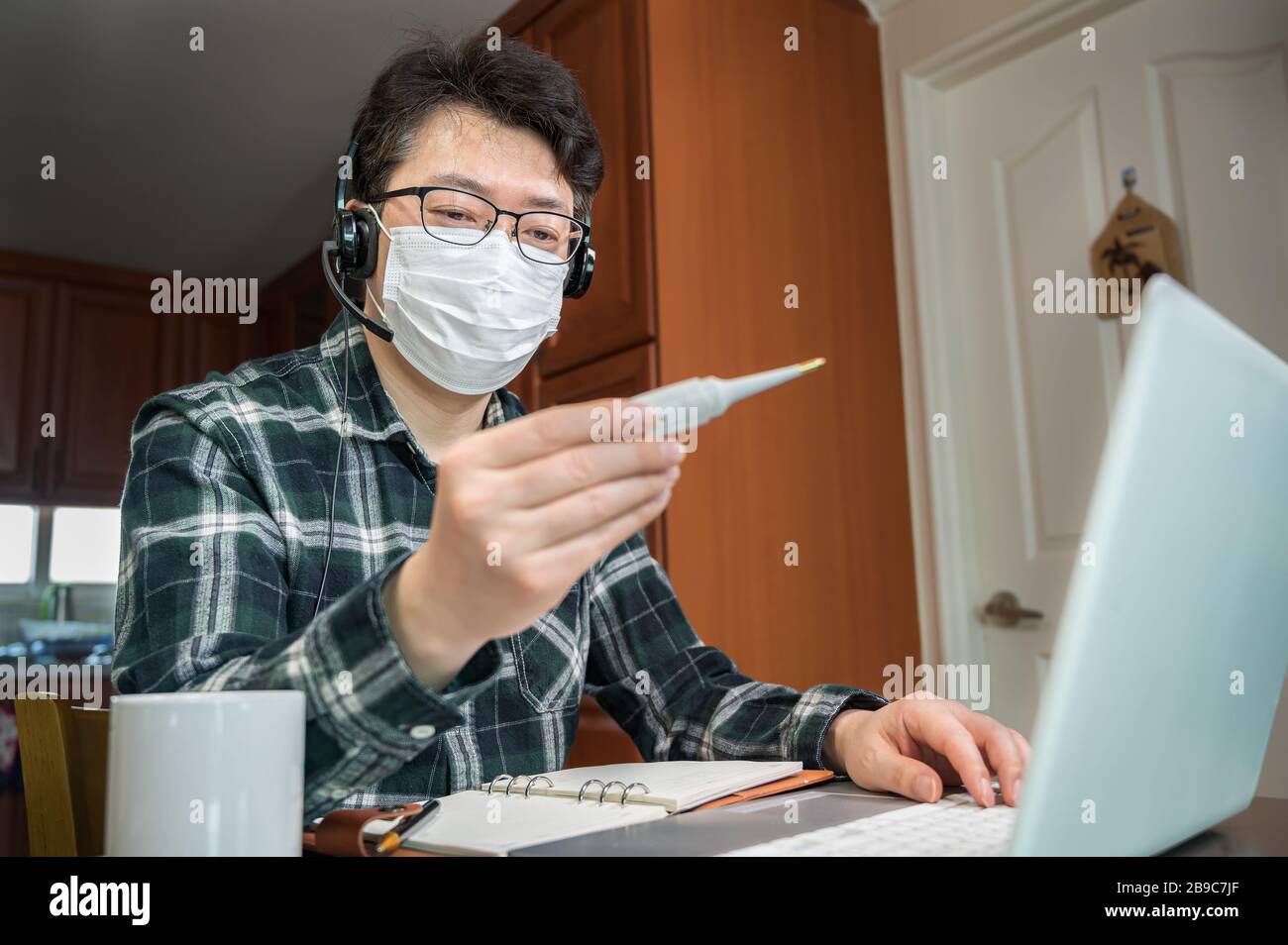 Ein asiatischer Mann, der aufgrund einer massiven Pandemie selbst isoliert und von zu Hause aus arbeitet. Ein Mann, der ein Thermometer hält, um die Körpertemperatur zu messen. Stockfoto