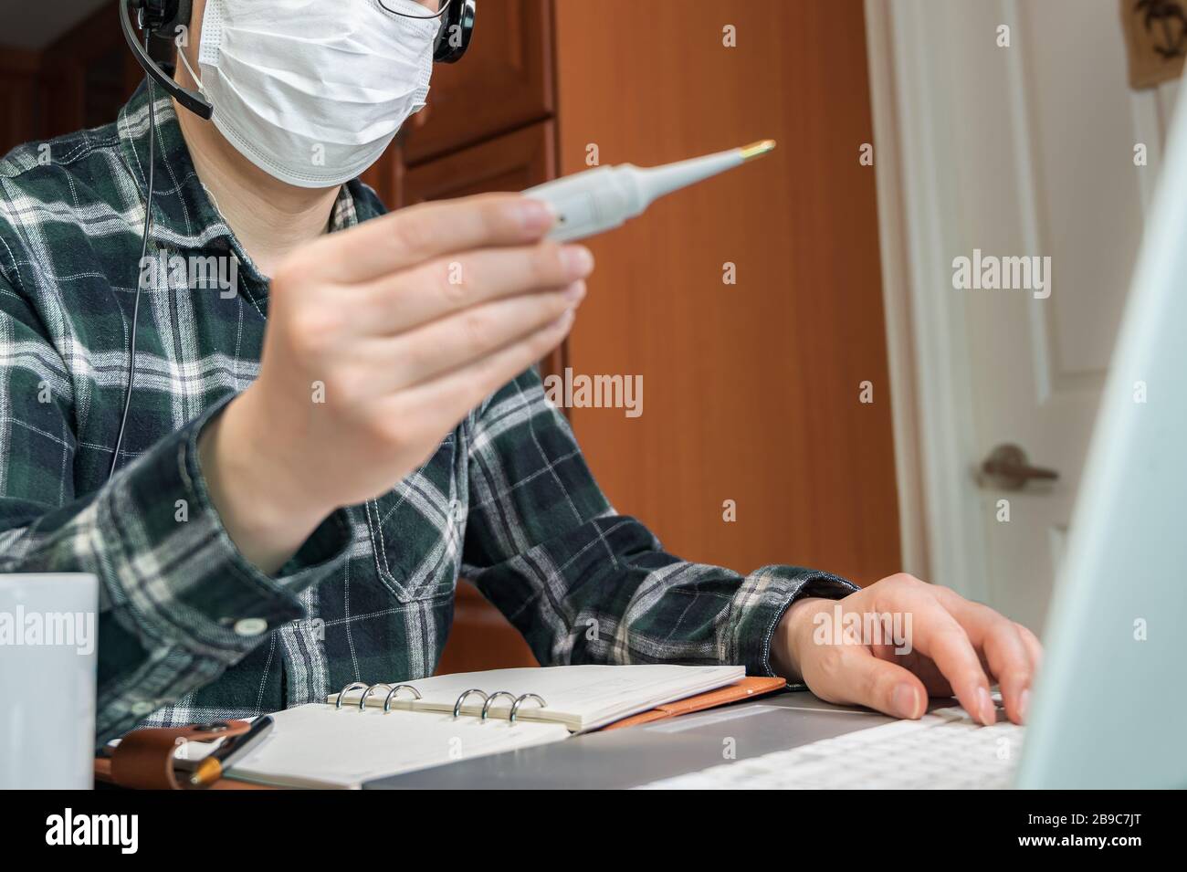 Ein asiatischer Mann, der aufgrund einer massiven Pandemie selbst isoliert und von zu Hause aus arbeitet. Ein Mann, der ein Thermometer hält, um die Körpertemperatur zu messen. Stockfoto