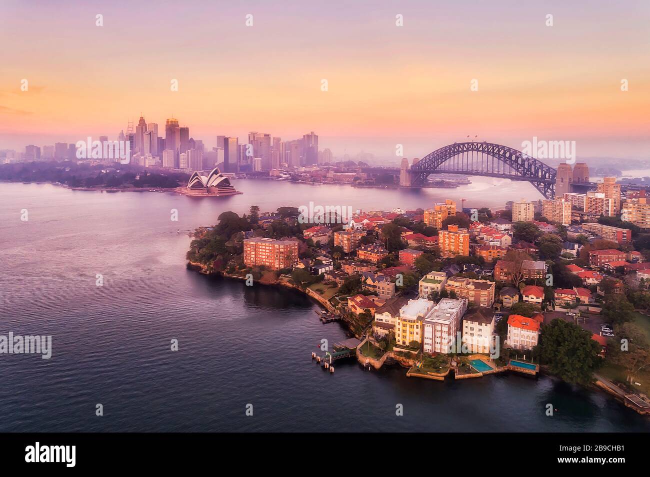 Hafen von Sydney und wichtige Wahrzeichen der Stadt am Wasser rund um die Sydney Harbour Bridge im Luftbild bei rosafarbenem Sonnenaufgang. Stockfoto