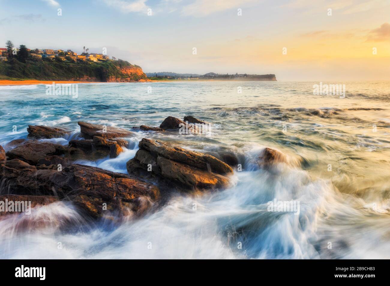 Der hohe Schlüssel der schnellen Wellen, die bei Sonnenaufgang über Sandsteinfelsen am Warriewood Beach in Sydney fließen. Stockfoto
