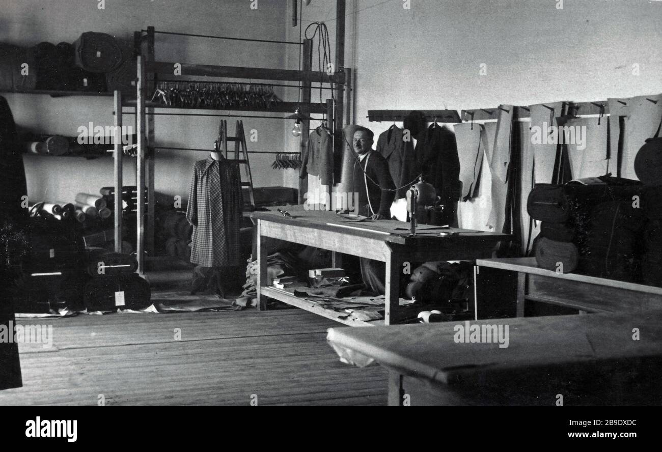 Ein Schneider steht in einem großen Raum hinter seinem Schneidetisch, der  von vielen Ballen aus Stoff umgeben ist. Das Bild wurde 1930 an einem  unbekannten Ort aufgenommen. [Automatisierte Übersetzung] Stockfotografie -  Alamy