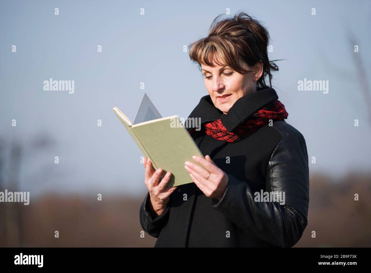 Eine Frau andere altert in der schwarzen Lederjacke ein Buch lesend vor unerscharfen Gerichtettern Hintergrund. Stockfoto