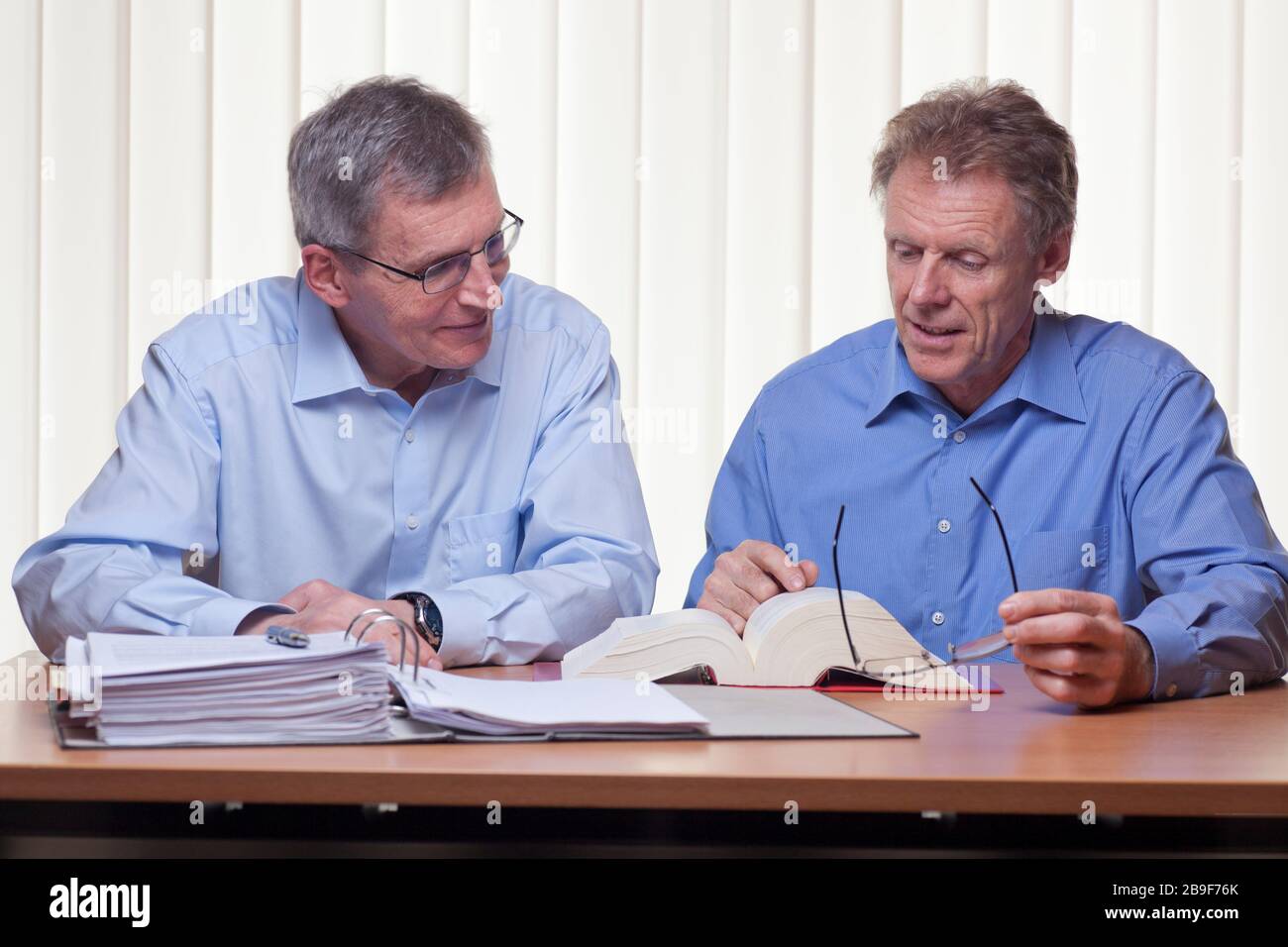 Zwei reife Geschäftsleute oder Partner lesen sich in einem Buch wieder, das an einem Schreibtisch mit Ordner und Buch sitzt - heller Hintergrund Stockfoto