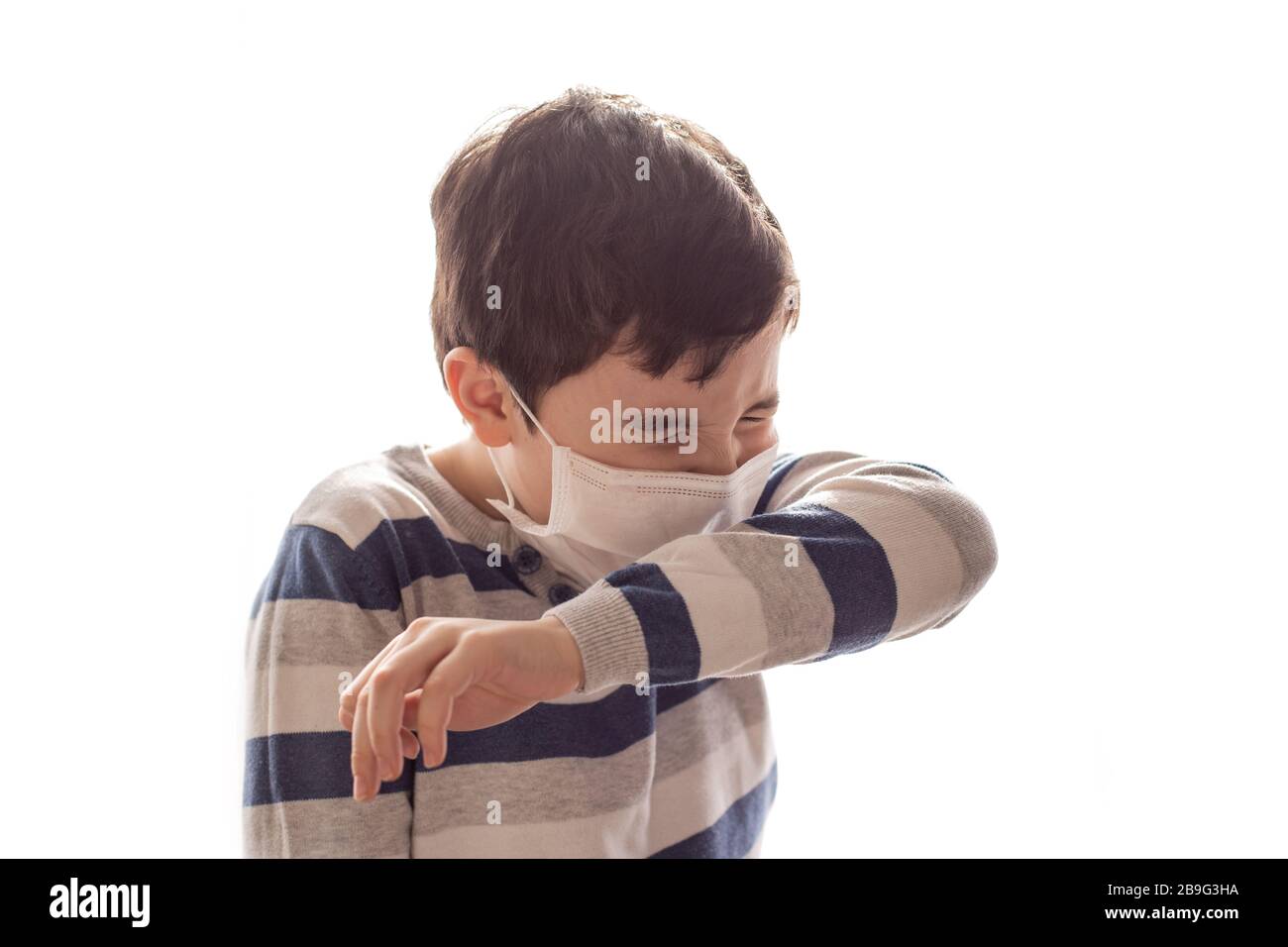 Ein Junge mit gequetteten Augen und einer weißen medizinischen Maske niest oder hustet in seinen Ellenbogen. Weißer Hintergrund. Kopierbereich. Influenza, Virus und Epidemie, coron Stockfoto