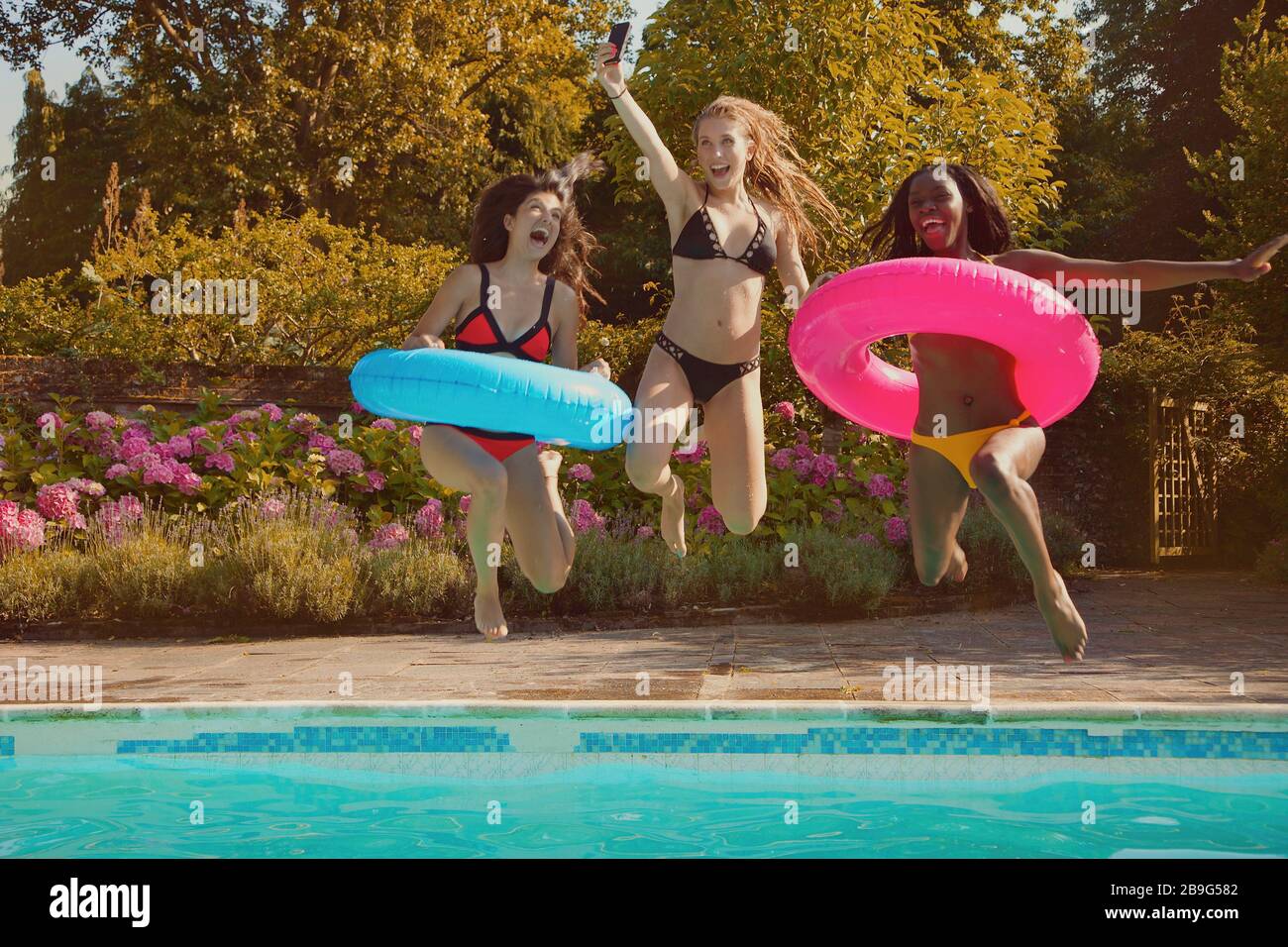 Verspielte Teenager-Freundinnen springen in den sonnigen Sommer-Swimmingpool Stockfoto