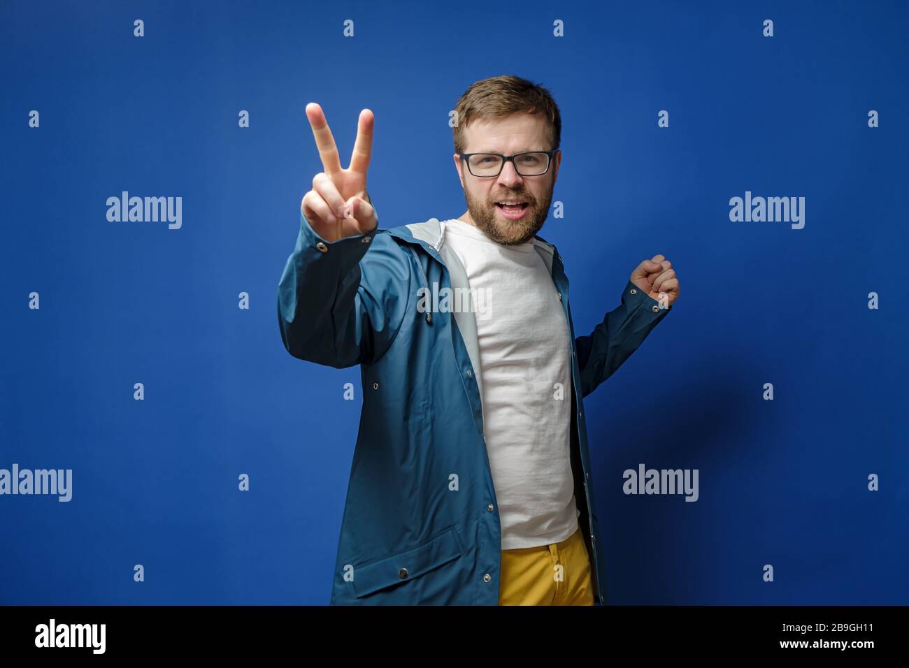 Kaukasischer, bärtiger Mann mit Brille und Regenmantel macht ein V-Zeichen mit Fingern und blickt auf die Kamera, auf blauem Hintergrund. Stockfoto
