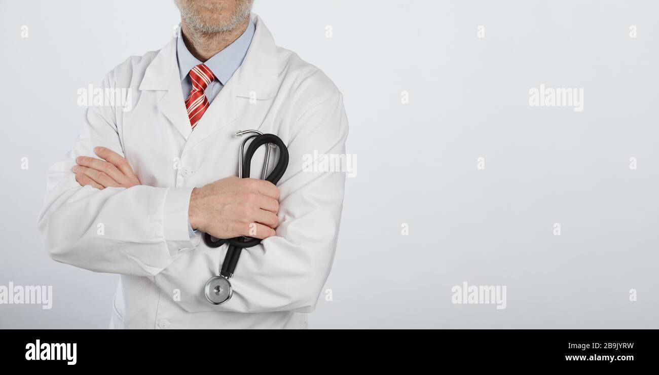 Stethoskop in den Händen eines bärtigen Arztes mit gekreuzten Armen. Arzt in einem blauen Hemd, roter Krawatte und weißem medizinischen Mantel Stockfoto