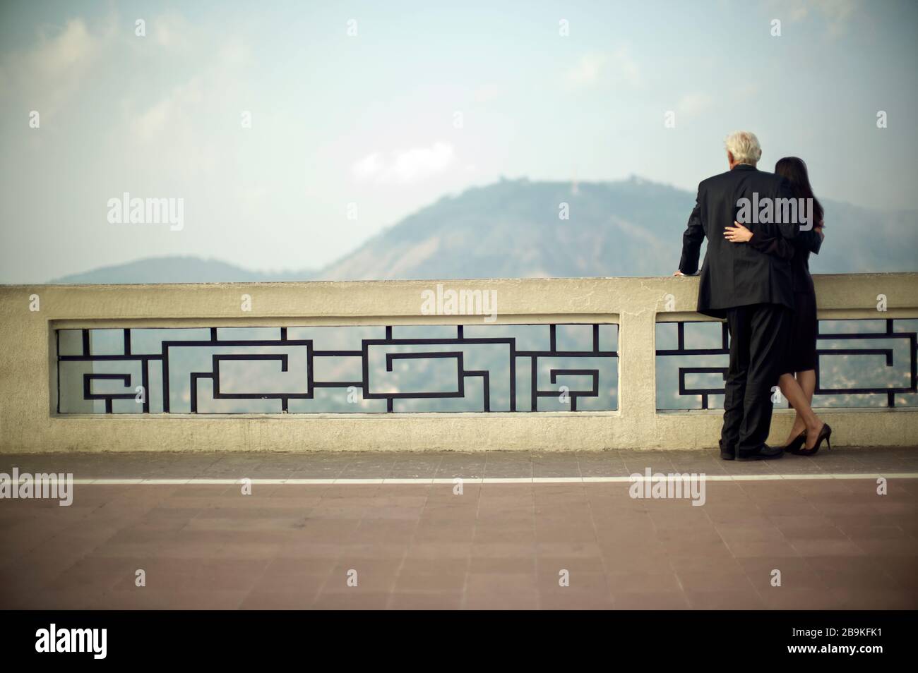 Buisnessman und Geschäftsfrau stehen mit Armen um sich herum, während sie von einem Balkon aus einen Blick auf die Hügel werfen. Stockfoto