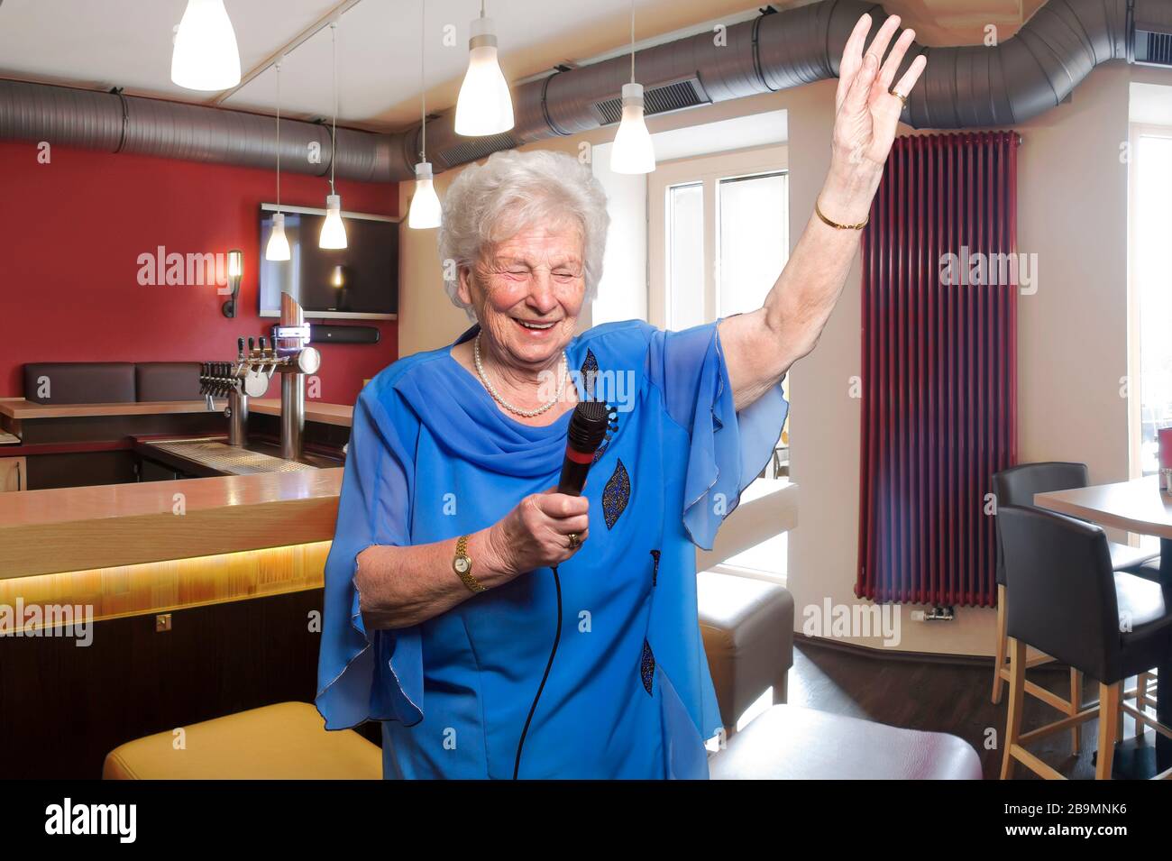 Die 80-jährige Frau singt Karaoke in einer Bar. Barhocker und Zähler im Hintergrund. Sie schafft eine gute Atmosphäre auf einer Party. Stockfoto