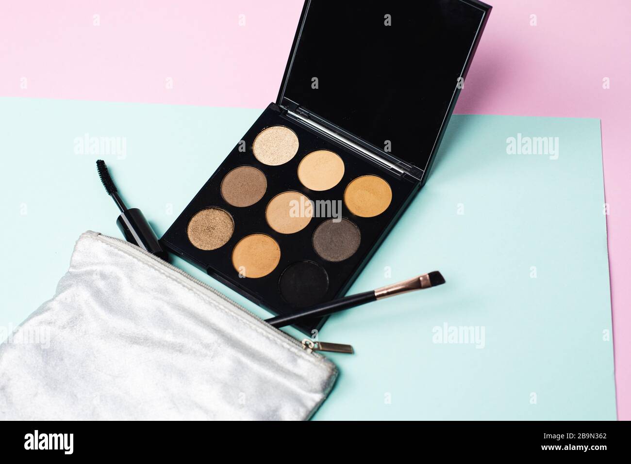 Eine kleine silberne Kosmetiktasche mit einer großen schwarzen Palette neutraler Töne, einer Mascara-Bürste und einer Bürste zum Auftragen von Kosmetika auf Pastellgrund. Das Konzept der schnellen täglichen Make-up. Stockfoto