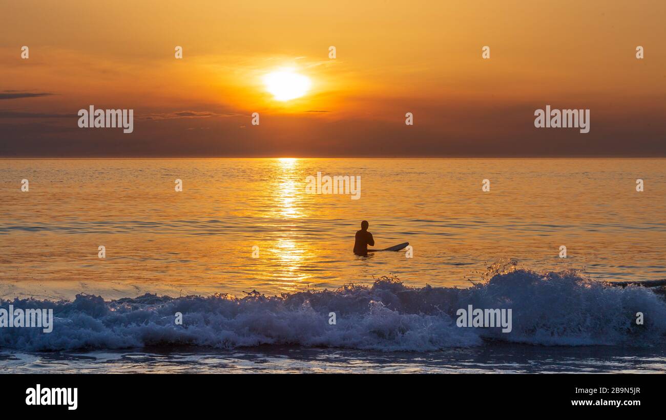 Ein Surfer, der auf ihrem Surfbrett sitzt, wartet im Wasser, um eine letzte Welle zu fangen, bevor die Sonne untergeht Stockfoto