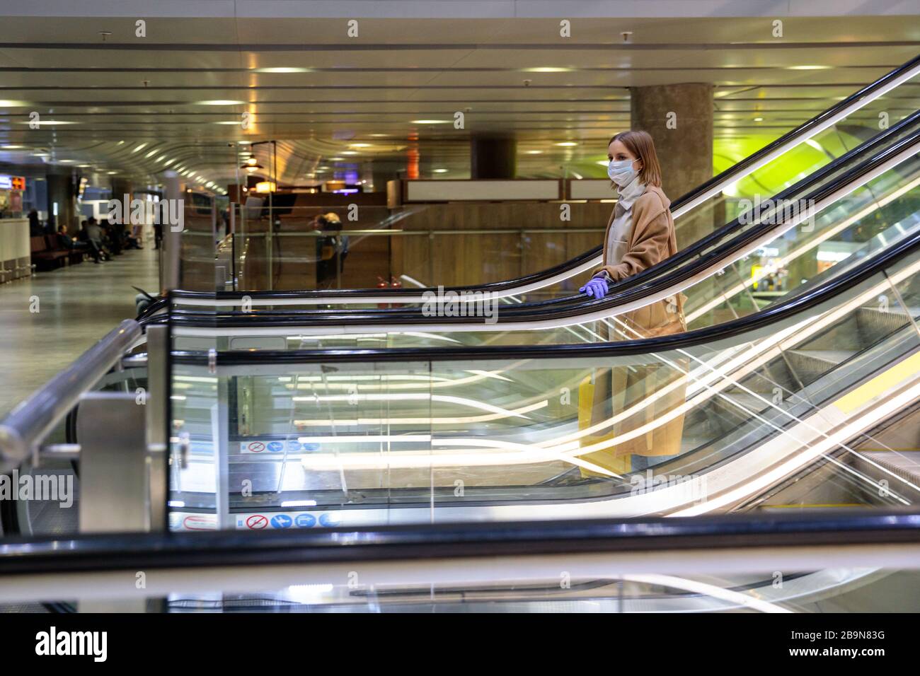 Frau mit gelbem Gepäck steht auf Rolltreppe, hält Rolltreppen-Handlauf im Flughafenterminal durch Latexhandschuhe, um sich vor Kontakt mit w zu schützen Stockfoto