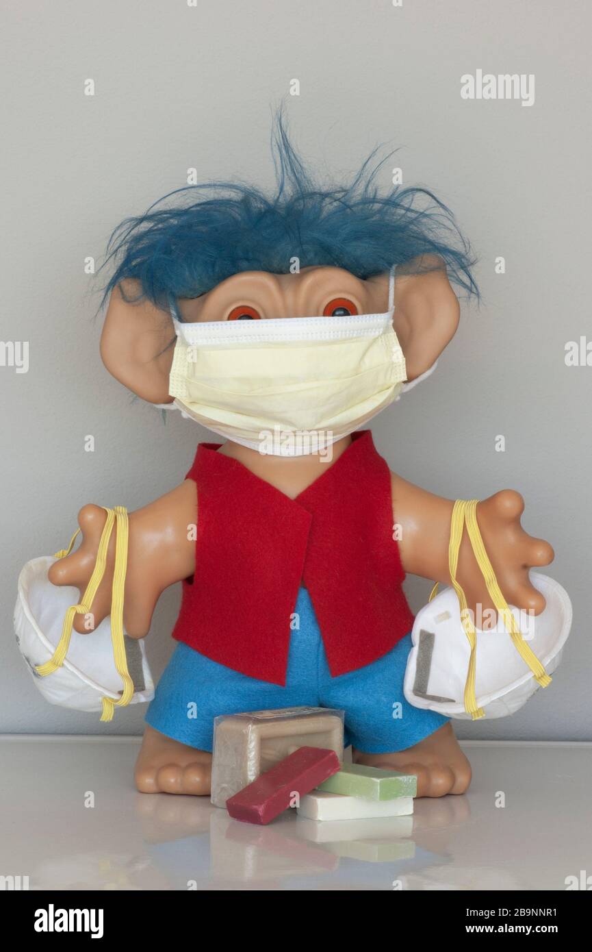 Troll-Kind trägt medizinische Maske und trägt mehr Masken. Seifenbarren befinden sich zu Füßen des Trolls. Eine Warnung zur Vorbeugung gegen COVID-19-Viren. Stockfoto