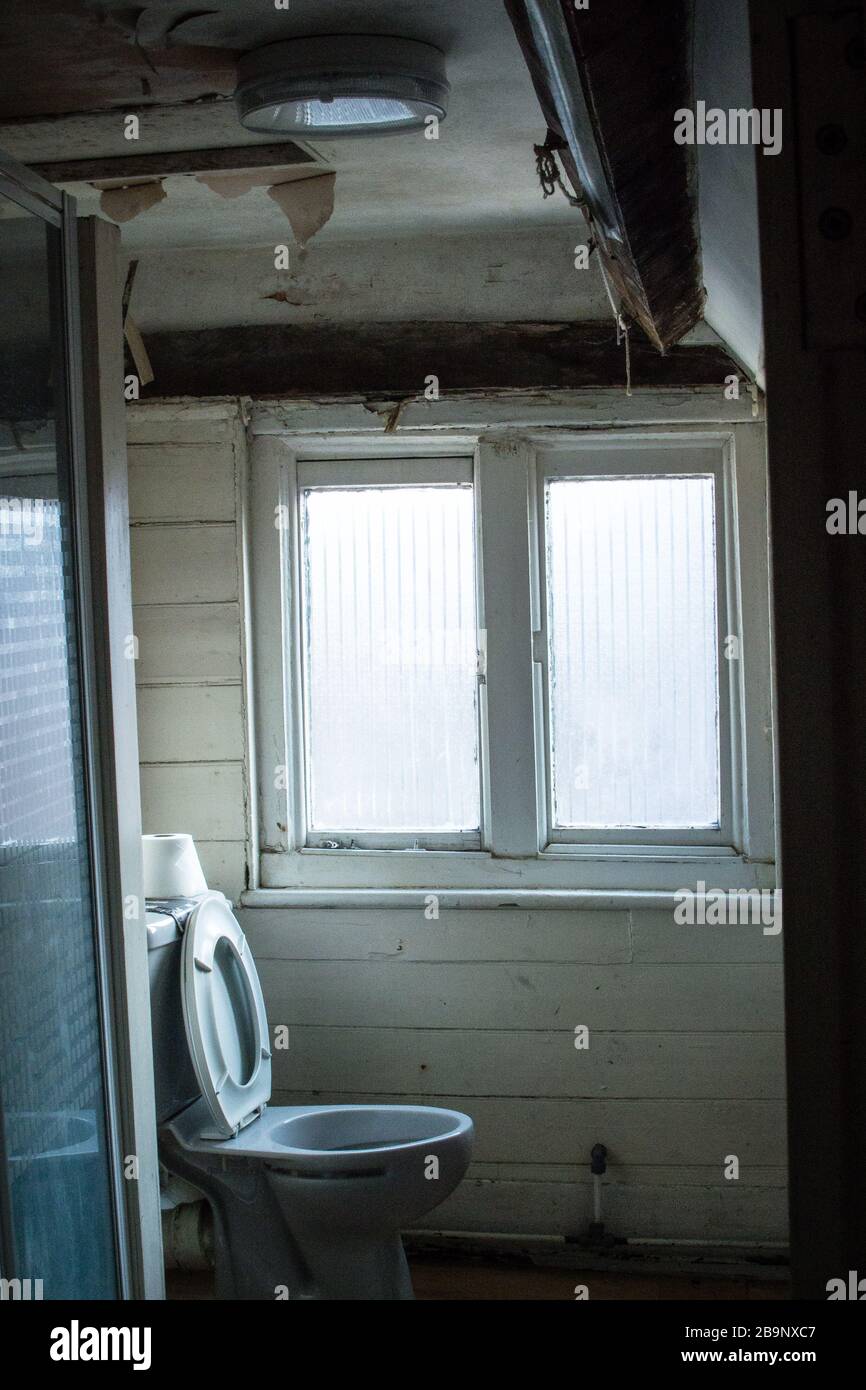 Toilette in einem verlassenen Haus. Die Decke fällt herunter. Toilettensitz nach oben. Toilettenpapier auf der Zisterne. Holzlatten Wände. Die Tapete blättert ab. Stockfoto